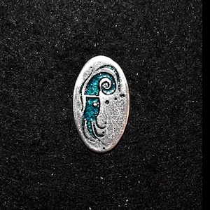 Op de afbeelding: Een zilveren broche met een turquoiseblauw ontwerp van een gestileerd zeedier. De broche is ovaalvormig en heeft een getextureerd oppervlak.