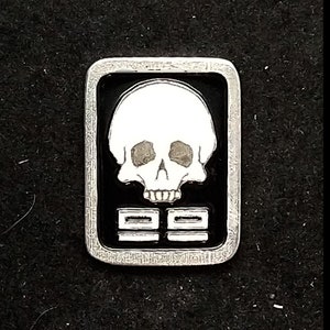 Könnte beinhalten: Eine silberfarbene Metallstifte mit schwarzem Hintergrund und einem weißen Totenkopfdesign. Der Schädel hat zwei Augenhöhlen und einen Mund mit Zähnen. Die Zahl "99" ist in weiß unter dem Schädel gedruckt.