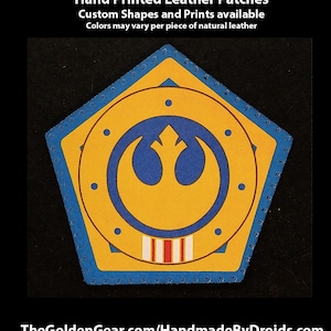 May include: A yellow and blue leather patch with a Rebel Alliance symbol in the centre. The patch is shaped like a pentagon with a blue border and stitching. The text "TheGoldenGear.com/HandmadeByDroids.com" is at the bottom of the image.