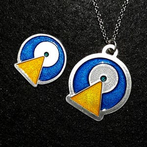 Peut inclure: Un collier en argent avec un pendentif bleu, blanc et jaune. Le pendentif est un cercle avec un cercle plus petit à l'intérieur et un triangle jaune pointant vers le haut. Il y a un petit pendentif assorti.