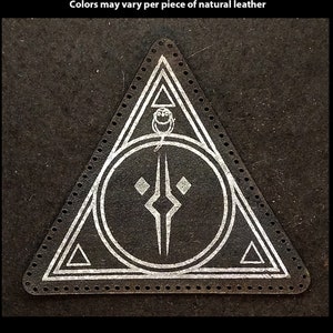 Op de afbeelding: Een handbedrukte leren patch in de vorm van een driehoek met een cirkel erin. De cirkel heeft een gestileerd ontwerp met een ruitvorm en een verticale lijn. De patch is op een zwarte achtergrond.