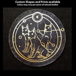 以下が含まれることがあります： 円形の黒い革のパッチで、金色のデザインが施されています。3匹のスタイリッシュな犬が幾何学模様の円の中に描かれています。画像の上部には「Hand Printed Leather Patches」というテキストがあります。最初のテキスト行の下には「Custom Shapes and Prints available」というテキストがあります。2行目のテキストの下には「Colors may vary per piece of natural leather」というテキストがあります。画像の下部には「TheGoldenGear.com/HandmadeByDroids.com」というテキストがあります。