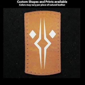 Könnte beinhalten: Ein braunes Lederpatch mit einem weißen geometrischen Design. Das Patch ist handgenäht an den Rändern. Der Text "Hand Printed Leather Patches" befindet sich oben im Bild. Der Text "Custom Shapes and Prints available" befindet sich unter der ersten Textzeile. Der Text "Colors may vary per piece of natural leather" befindet sich unter der zweiten Textzeile. Der Text "TheGoldenGear.com/HandmadeByDroids.com" befindet sich unten im Bild.
