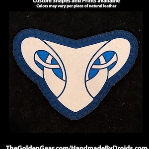 May include: A white leather patch with a blue outline in the shape of a stylized snake head with blue eyes. The patch is hand-printed and available in custom shapes and prints. "TheGoldenGear.com/HandmadeByDroids.com" is printed below the patch.