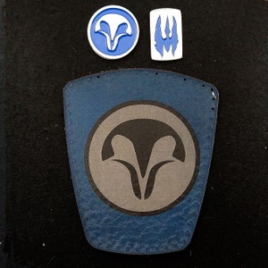 Puede incluir: Un parche de cuero azul con un borde cosido y un símbolo gris en forma de V estilizada dentro de un círculo.