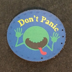 Peut inclure: Un patch rond en cuir bleu avec un personnage extraterrestre vert avec une langue qui sort. L'extraterrestre est entouré d'un cercle jaune avec le texte "Don't Panic".