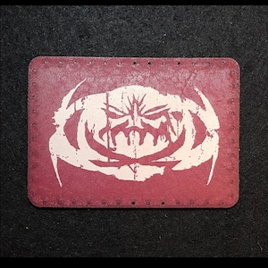 May include: A red leather patch with a white hand-printed design of a skull with a crown. The patch has a stitched border and is available in custom shapes and prints.