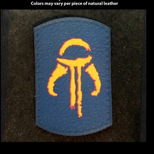 Könnte beinhalten: Ein blauer Leder-Patch mit einem gelben und orangefarbenen Symbol. Das Symbol ist eine stilisierte Darstellung eines mandalorianischen Symbols. Der Patch ist handgefertigt und in benutzerdefinierten Formen und Drucken erhältlich. TheGoldenGear.com/HandmadeByDroids.com
