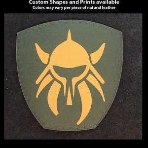 May include: A green leather patch with a yellow Viking helmet design. The patch is hand-printed and has a stitched edge. The text "TheGoldenGear.com/HandmadeByDroids.com" is printed below the patch.