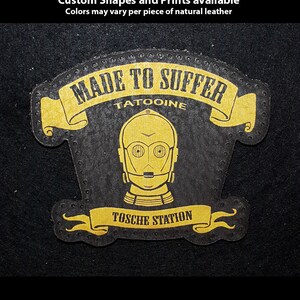 May include: Hand-printed leather patch featuring a gold C-3PO design with the text "MADE TO SUFFER" and "TOSCHE STATION" on a black background. The patch is custom-shaped and printed, with the text "TATOOINE" above the C-3PO image.