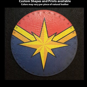 May include: A round leather patch with a yellow star and two yellow stripes on a red and blue background. The text "Hand Printed Leather Patches" is at the top of the image. The text "Custom Shapes and Prints available" is below the first line of text. The text "Colors may vary per piece of natural leather" is below the second line of text. The text "TheGoldenGear.com/HandmadeByDroids.com" is at the bottom of the image.