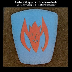 May include: A blue leather patch with a red and black stylized monster design. The patch is hand-printed and has a stitched edge.