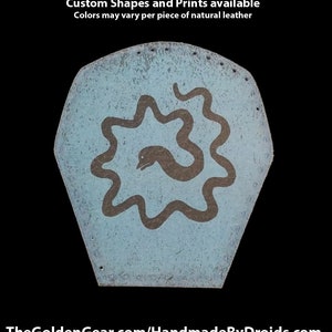 May include: A light blue leather patch with a hand-painted design of a snake curled around a star shape. The text "Hand Printed Leather Patches" is at the top of the image. The text "Custom Shapes and Prints available" is below the first line of text. The text "Colors may vary per piece of natural leather" is below the second line of text. The text "TheGoldenGear.com/HandmadeByDroids.com" is at the bottom of the image.