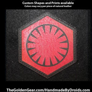 以下が含まれることがあります： 六角形の赤い革のパッチで、放射状の線を持つ円形のスタイリッシュなシンボルが描かれています。画像の上部に「Hand Printed Leather Patches」というテキストがあります。最初のテキスト行の下に「Custom Shapes and Prints available」というテキストがあります。2行目のテキストの下に「Colors may vary per piece of natural leather」というテキストがあります。画像の下部に「TheGoldenGear.com/HandmadeByDroids.com」というテキストがあります。
