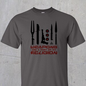 May include: A dark gray t-shirt with a black graphic of various weapons and the text "WEAPONS ARE A PART OF MY RELIGION" in red.