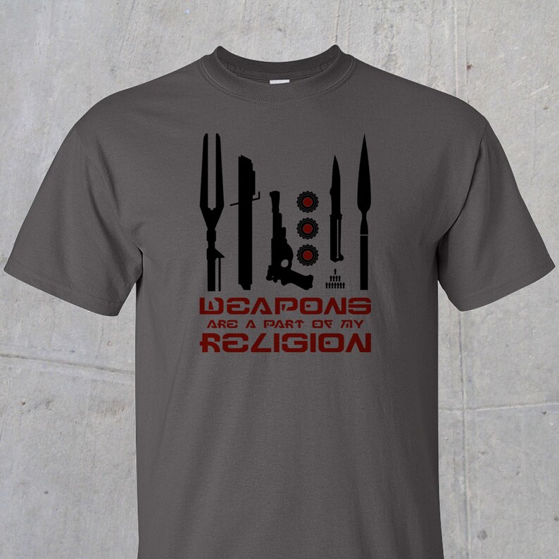 May include: A dark gray t-shirt with a black graphic of various weapons and the text "WEAPONS ARE A PART OF MY RELIGION" in red.