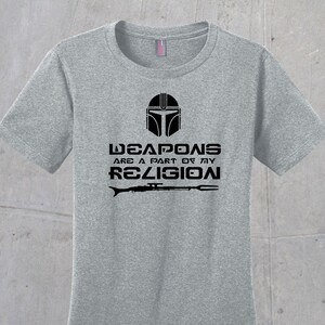 May include: Grey t-shirt featuring a black Mandalorian helmet graphic and the text "Weapons are a part of my religion" with a black rifle and spear graphic below.