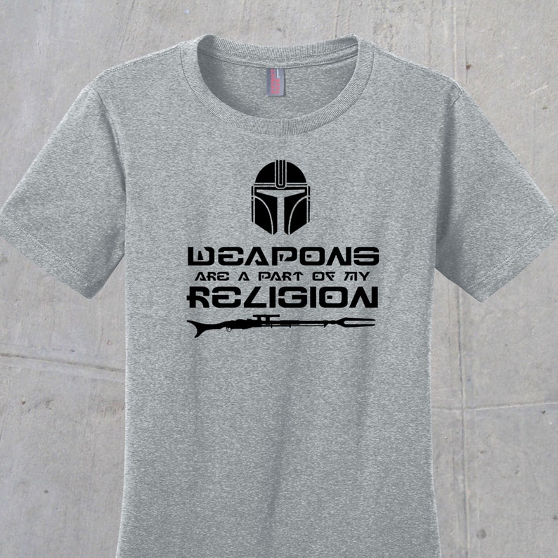 May include: Grey t-shirt featuring a black Mandalorian helmet graphic and the text "Weapons are a part of my religion" with a black rifle and spear graphic below.
