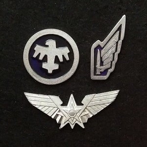 Op de afbeelding: Drie zilveren metalen pinnen met verschillende ontwerpen. De eerste pin is een cirkel met een blauwe achtergrond en een witte adelaar met een kruis in het midden. De tweede pin is een zilveren vleugel met een dolk in het midden. De derde pin is een zilveren vleugel met een ster in het midden.