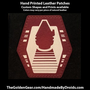 May include: A red leather patch with a white design of a spaceship. The patch is hand-printed and has a stitched edge. The text "Hand Printed Leather Patches" is at the top of the image. The text "Custom Shapes and Prints available" is below the first line of text. The text "Colors may vary per piece of natural leather" is below the second line of text. The text "TheGoldenGear.com/HandmadeByDroids.com" is at the bottom of the image.