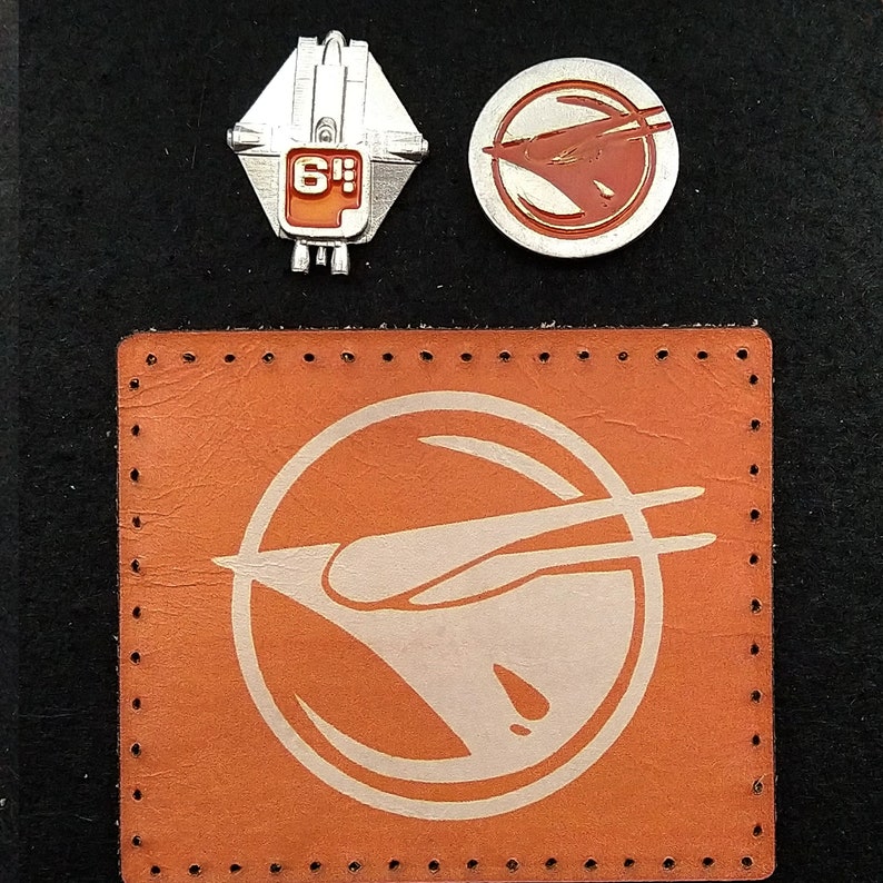 May include: A brown leather patch with a white stitched border and a white stylized image of a spaceship with the text "6'" inside a circle.