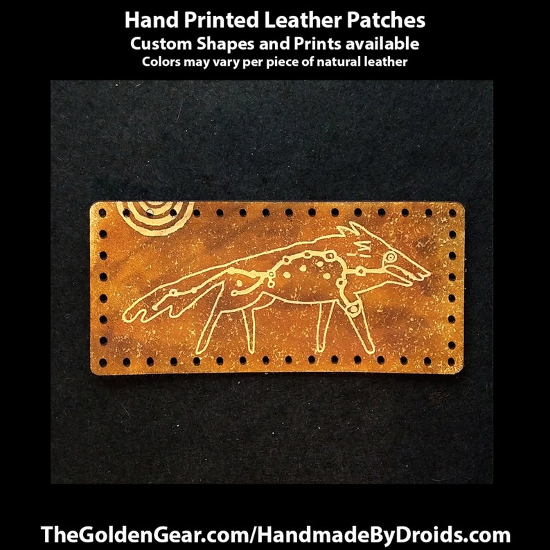 Pu&ograve; includere: Toppa in pelle stampata a mano con un disegno di lupo stilizzato in oro. La toppa rettangolare ha piccoli fori sui bordi. Il testo sulla toppa recita "Hand Printed Leather Patches" e "Custom Shapes and Prints available".