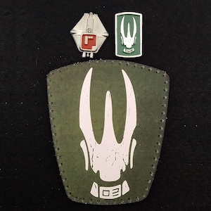 May include: A green patch with a white symbol and the text "02" on a brown leather patch with stitching around the edges.