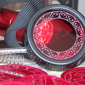 May include: A pair of black steampunk goggles with red lenses and a decorative design. The goggles are attached to a black strap and have a red light source inside.