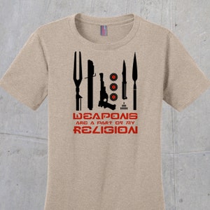 May include: A tan t-shirt with a black graphic of various weapons, including a fork, a lightsaber, a pistol, a knife, and a spear. The text "WEAPONS ARE A PART OF MY RELIGION" is printed in red below the graphic.