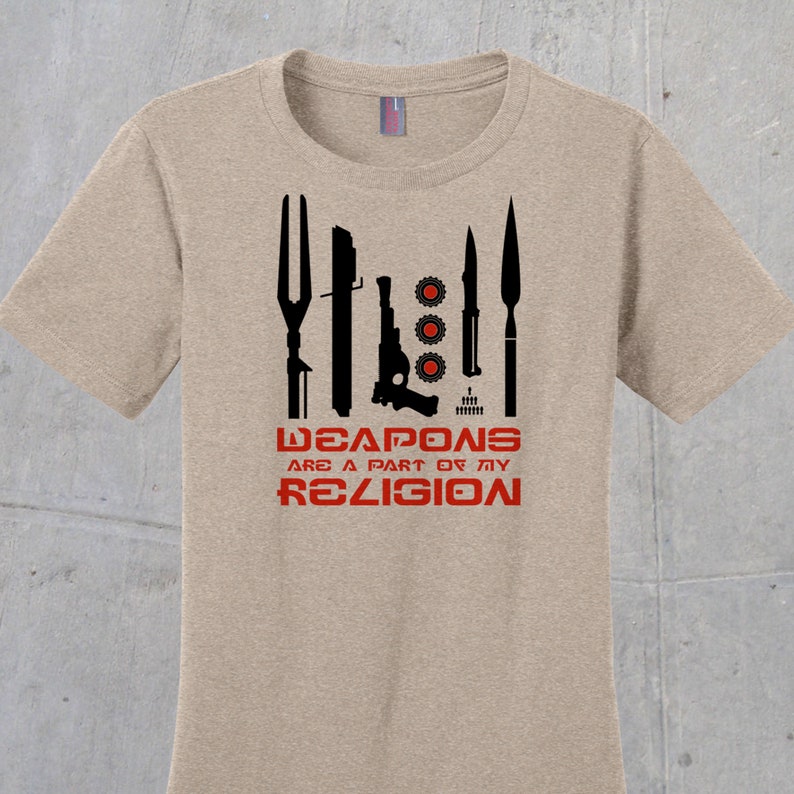May include: A tan t-shirt with a black graphic of various weapons, including a fork, a lightsaber, a pistol, a knife, and a spear. The text "WEAPONS ARE A PART OF MY RELIGION" is printed in red below the graphic.