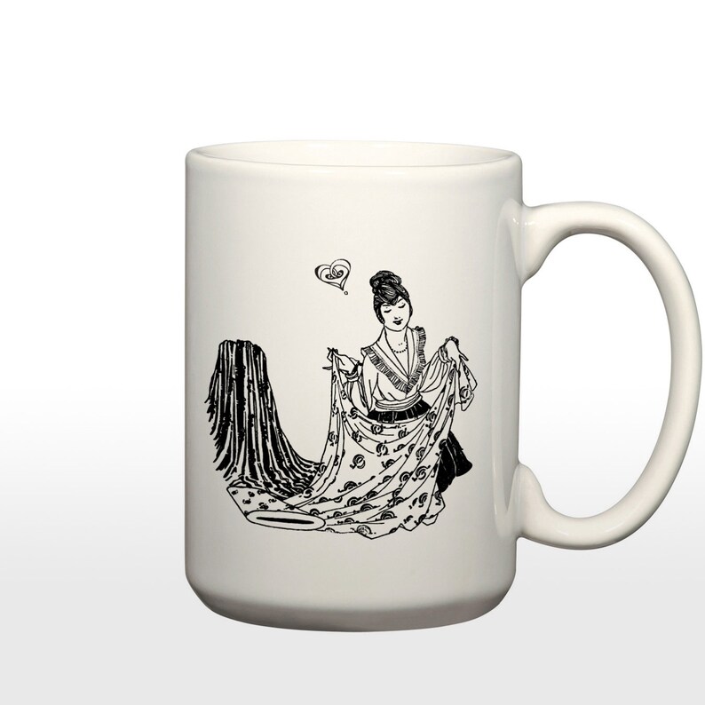 May include: White ceramic mug with a black and white illustration of a woman holding a patterned fabric. The woman is wearing a dress and has her hair pulled back. There is a heart above her head.