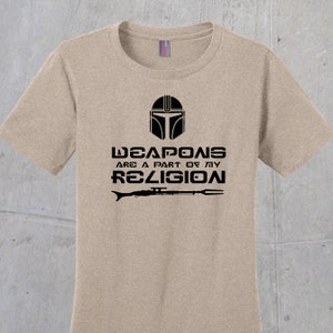 May include: A tan t-shirt with a black graphic of a Mandalorian helmet and the text "Weapons are a part of my religion" with a rifle and spear underneath.