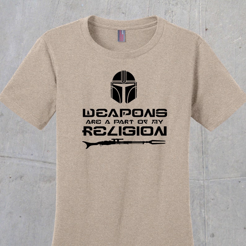 May include: A tan t-shirt with a black graphic of a Mandalorian helmet and the text "Weapons are a part of my religion" with a rifle and spear underneath.