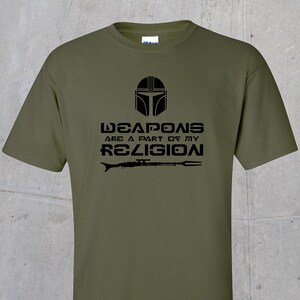 May include: A green t-shirt featuring a black graphic of a Mandalorian helmet and the text "Weapons are a part of my religion".