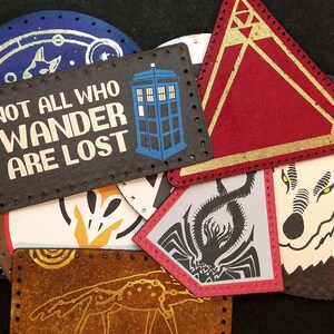 Pode incluir: Uma cole&ccedil;&atilde;o de nove patches de tecido coloridos com v&aacute;rios designs, incluindo um patch azul e prateado com a cita&ccedil;&atilde;o "Not all who wander are lost", um patch triangular vermelho e dourado, um patch azul e branco com um TARDIS e um patch marrom com uma ilustra&ccedil;&atilde;o de lobo branco.