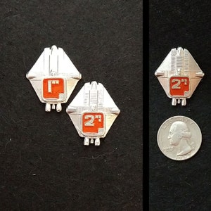 May include: Two silver metal badges with red numbers and the text "2" and "1" on them. The badges are shaped like a diamond with rounded corners and have a design that resembles a spaceship.