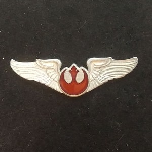 May include: A silver-coloured pin featuring a red and white emblem with wing details. The pin has a symmetrical design with a central emblem and wings extending outwards. The wings have detailed feather patterns.