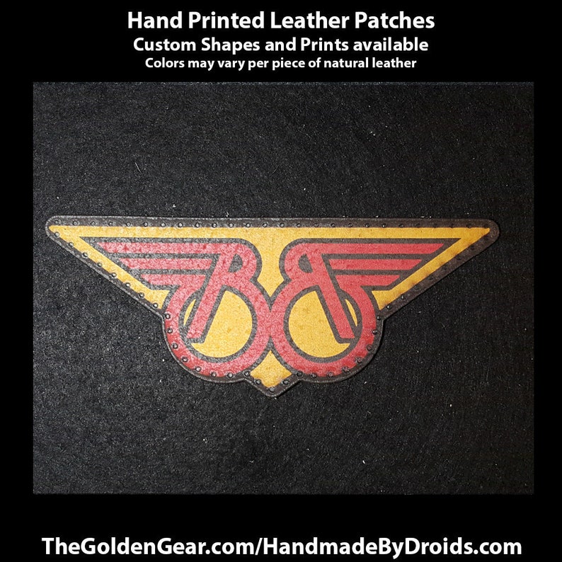 May include: A hand-painted leather patch with a red and yellow logo of two wings and the letters "8&" in the center. The patch is on a black background. TheGoldenGear.com/HandmadeByDroids.com