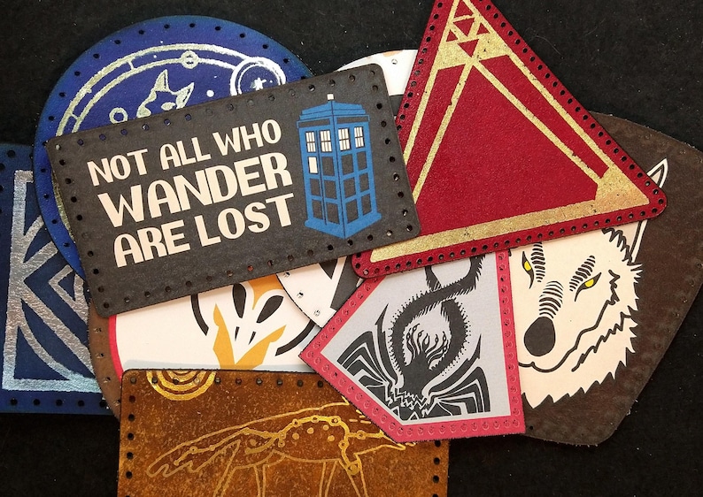 Puede incluir: Una colecci&oacute;n de nueve parches de tela coloridos con varios dise&ntilde;os, incluyendo un parche azul y plateado con la cita "Not all who wander are lost", un parche triangular rojo y dorado, un parche azul y blanco con un TARDIS y un parche marr&oacute;n con una ilustraci&oacute;n de lobo blanco.