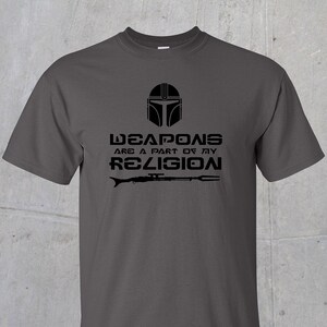 May include: A dark grey t-shirt with a black graphic of a Mandalorian helmet and the text "Weapons are a part of my religion".