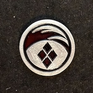 May include: A silver pin with a black and red design. The design features a stylized diamond shape with a wave-like shape around it.