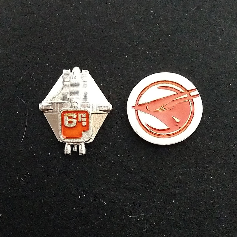May include: Two silver metal pins. One pin is shaped like a spaceship with a red number "6" on it. The other pin is round with a red spaceship design inside a white circle.