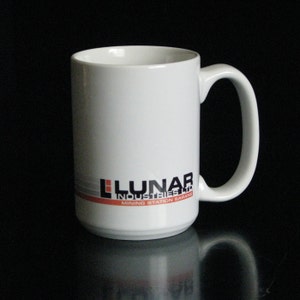 May include: White ceramic mug with a curved handle. The mug features the text "LUNAR INDUSTRIES LTD MINING STATION GARANG" in black, red, and gray. The mug is on a black surface.