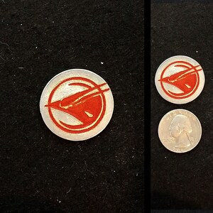 May include: A silver-colored metal token with a red stylized bird design. The bird is facing left and has a long, thin beak. The token is about the size of a US quarter.
