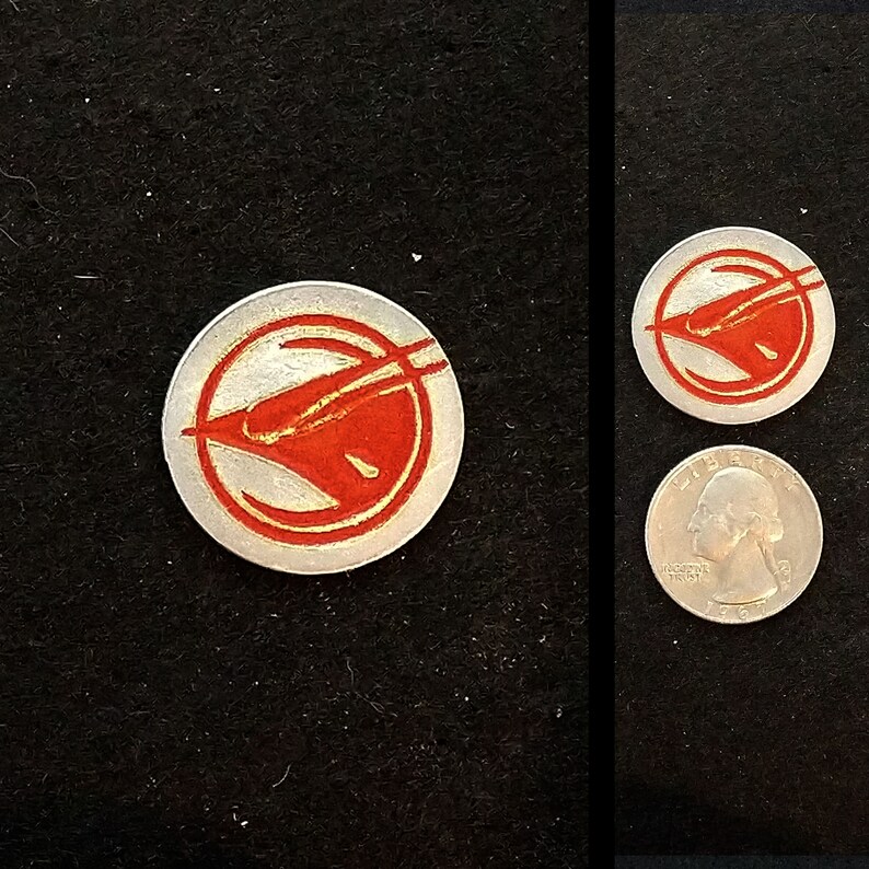 May include: A silver-colored metal token with a red stylized bird design. The bird is facing left and has a long, thin beak. The token is about the size of a US quarter.