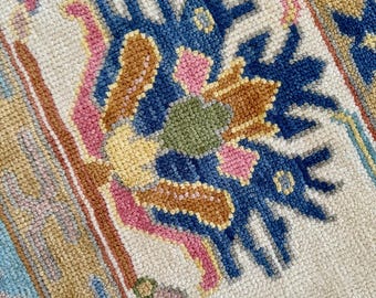 May include: Close-up of a section of a rug with a cream-colored background. The rug features a floral design in shades of blue, pink, brown, green, and yellow. The rug has a textured appearance.