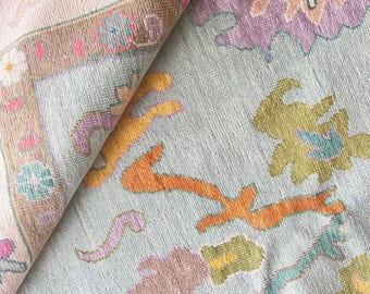 Può includere: Un tappeto piegato con sfondo azzurro e un motivo floreale dettagliato nei toni dell'arancione, viola, verde e rosa. Il tappeto ha un bordo color crema con finiture marroni e verde acqua.