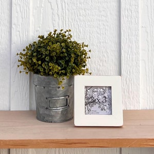 Könnte beinhalten: Weißer Bilderrahmen mit einem weißen Passepartout und einer Öffnung von 7,6 x 7,6 cm. Der Rahmen steht auf einem Holzregal mit einer kleinen Topfpflanze davor.