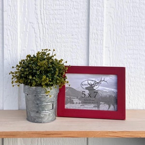 May include: A red 5x7 picture frame made of solid wood. The frame is handmade in the USA and features a rustic design with a deer silhouette. The frame is sitting on a wooden shelf with a small potted plant in front of it.