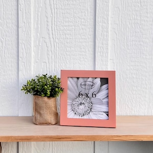 May include: A light pink wooden picture frame with a white flower inside. The frame is 6x6 inches and says "Handmade in USA Solid Wood" on the bottom.